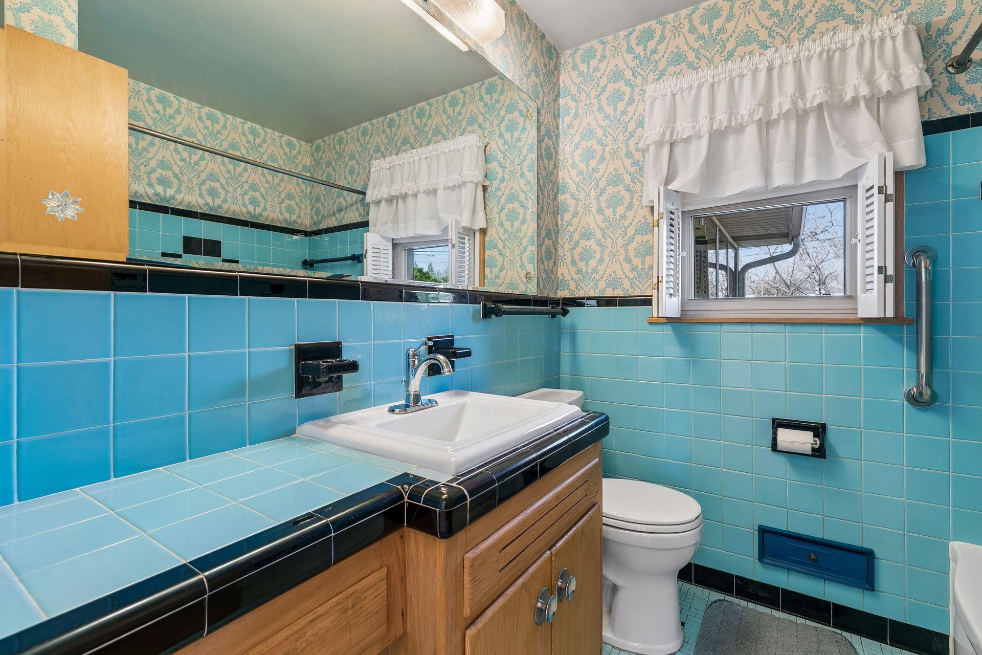 Main floor bathroom showcasing cool blue tile, a spacious mirror, wall mounted lighting, vanity sink, window, and a full tub/shower.