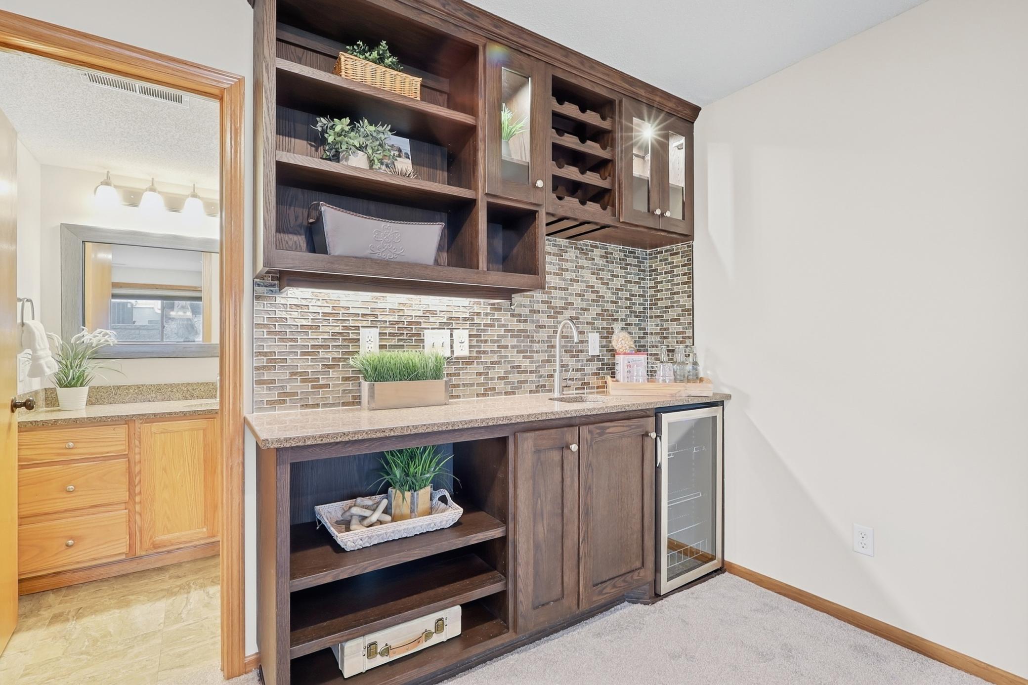 Wet bar with granite counters, tiled backsplash, beverage fridge, lighted glass cabinets and sink.