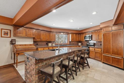 âSpacious kitchen with warm wood cabinetry, granite counters, a large island with seating, brick accents, stainless appliances, and a wide window bringing in natural light.â