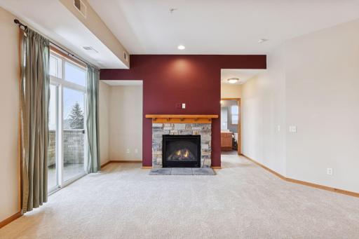 Bright living room with plush carpeting, stone-surround gas fireplace, and sliding glass door leading to a private balcony with scenic views of the nearby path.