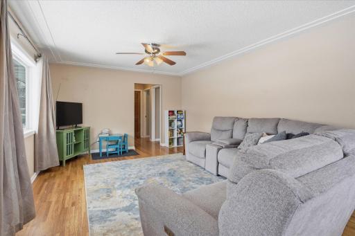 Bright living room with large picture window, ceiling fan, and warm hardwood flooring. Layout offers easy access to bedrooms while maintaining an open feel.
