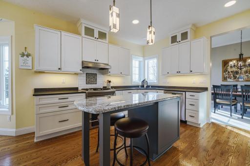 The furniture-style island has a granite top and space for seating and storage. Two pendant lights hang above the island. The primary countertops are illuminated by undercabinet lighting.