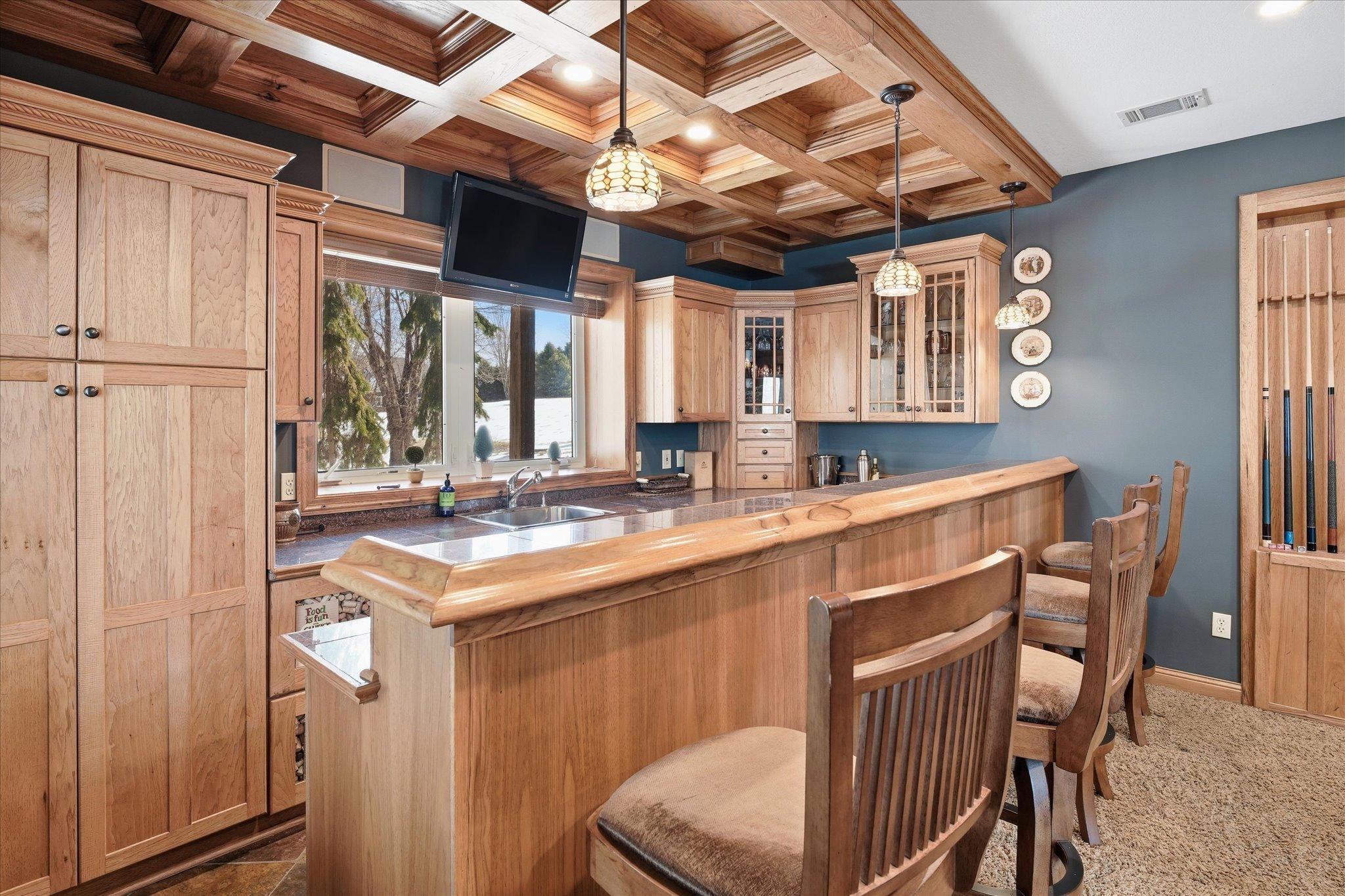 Stunning lower-level bar area showcasing exceptional craftsmanship with rich custom wood cabinetry, a beautifully coffered wood ceiling, granite countertops, and an expansive bar perfect for entertaining.
