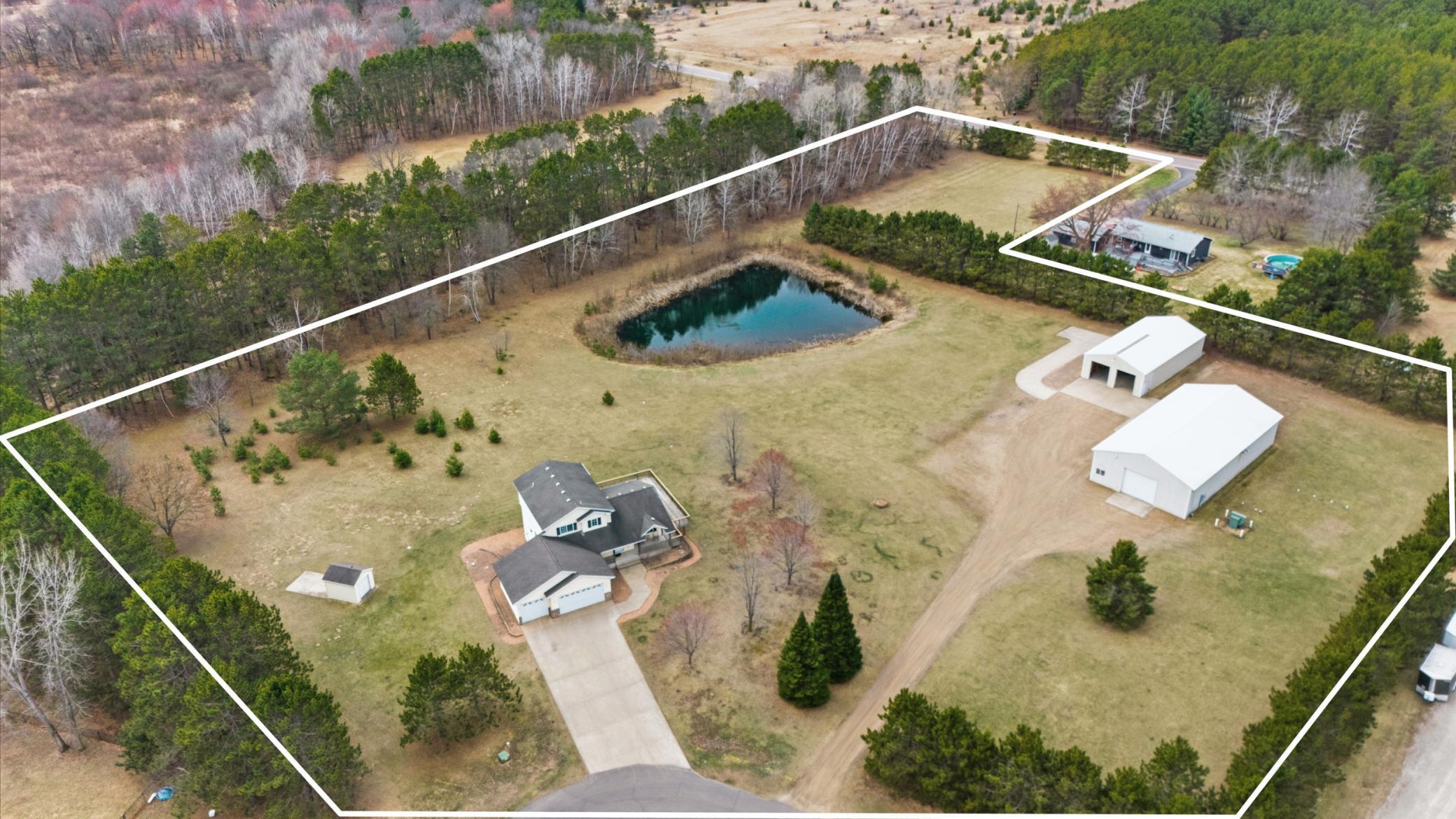 Aerial overview highlights the full property layout including home, pond, acreage, and both impressive outbuildings.