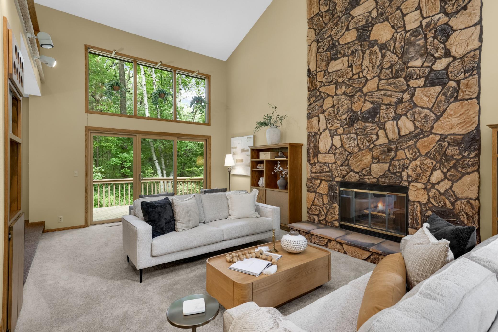The dramatic two-story living room features a striking floor-to-ceiling stone fireplace and expansive windows overlooking the wooded backyard.