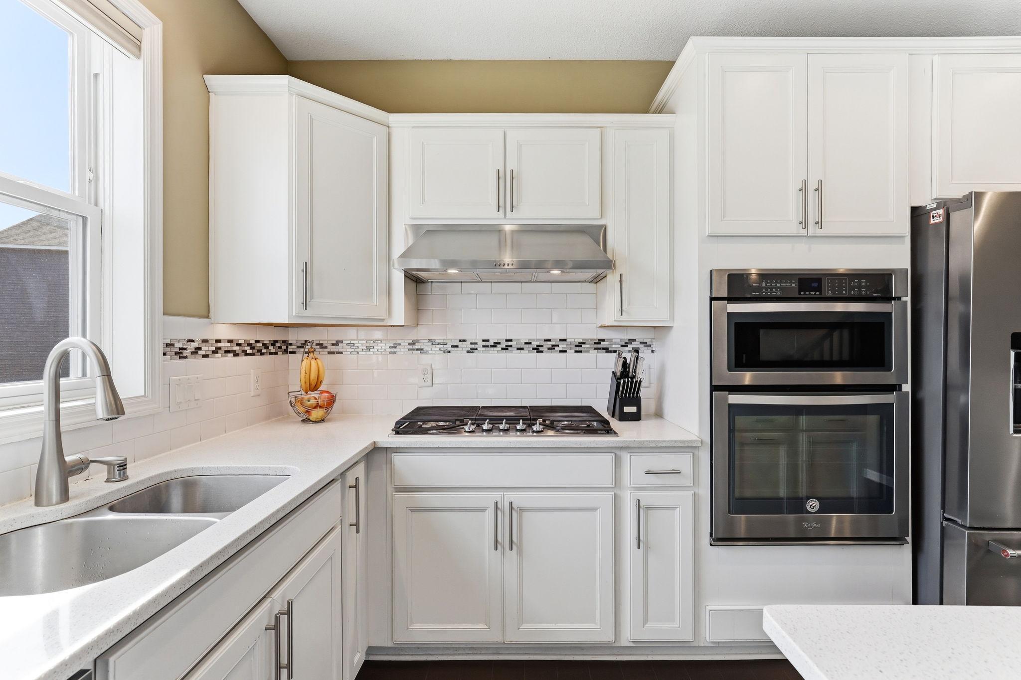 Double-wall ovens and an undermount sink below a light-filled window anchor the kitchen's working wall. Quartz counters, subway tile backsplash, and custom white cabinetry complete a workspace built to perform.
