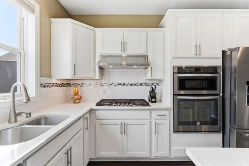 Double-wall ovens and an undermount sink below a light-filled window anchor the kitchen's working wall. Quartz counters, subway tile backsplash, and custom white cabinetry complete a workspace built to perform.