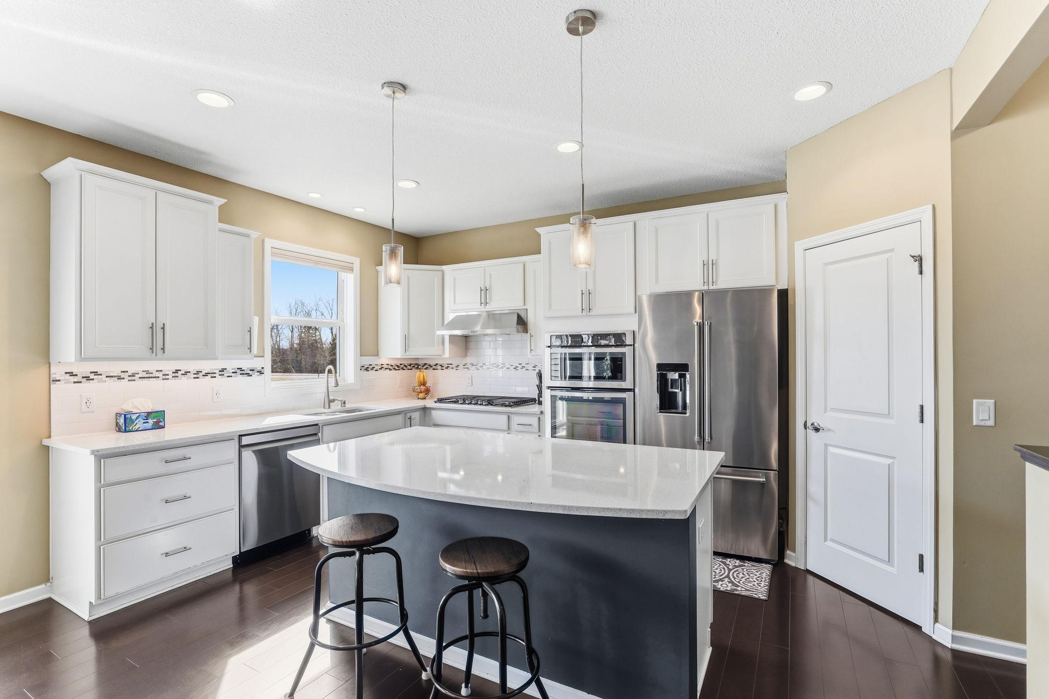 Quartz counters, a 5-burner gas cooktop, double-wall ovens, and a stainless steel French-door refrigerator deliver serious cooking capability. A window above the sink brings light directly to the workspace.