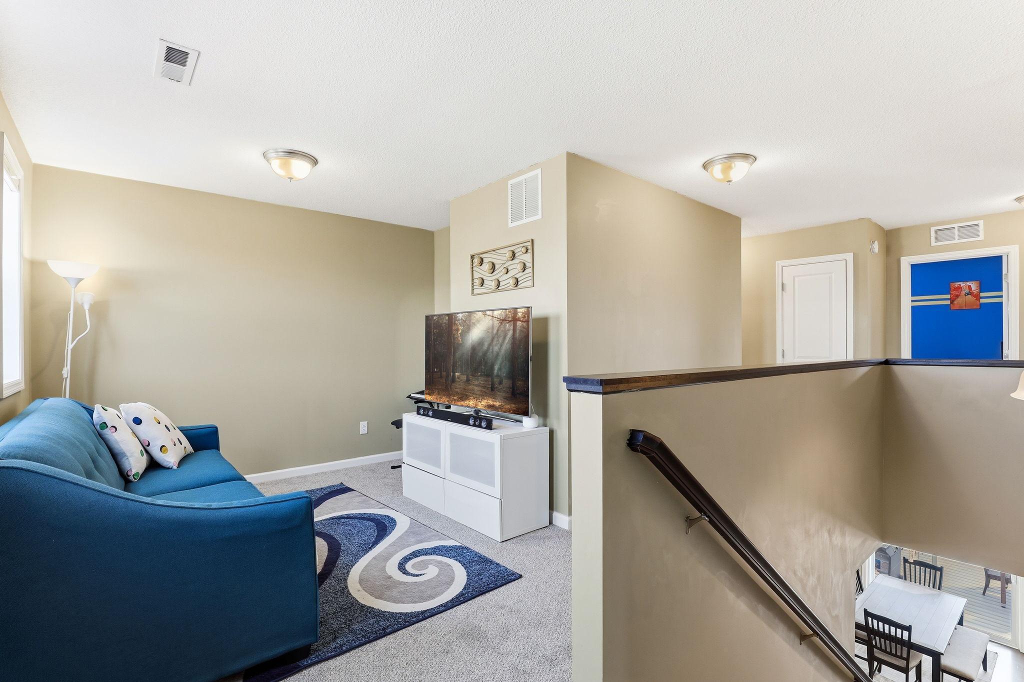 At the top of the stairs, a carpeted flex space serves as a second media room, play area, or homework hub, with an open railing overlooking the main level and doors to the bedrooms beyond.