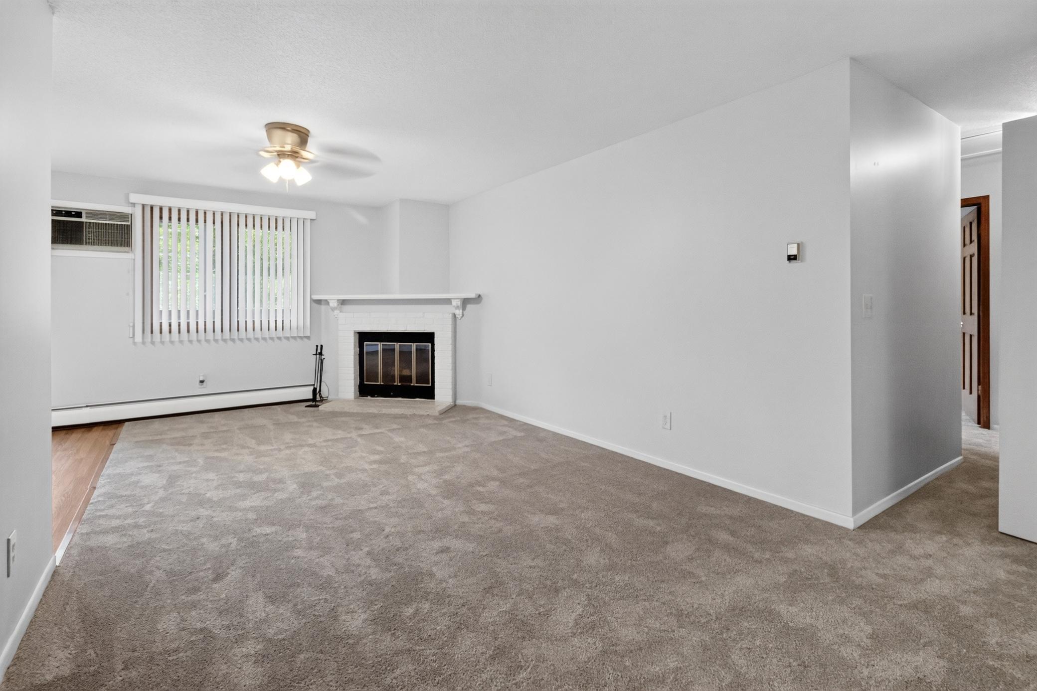 Spacious and sunlit living room features an inviting brick-wrapped fireplace with a white mantel, comfortable carpeting, a ceiling fan, and a view into the adjoining dining room.