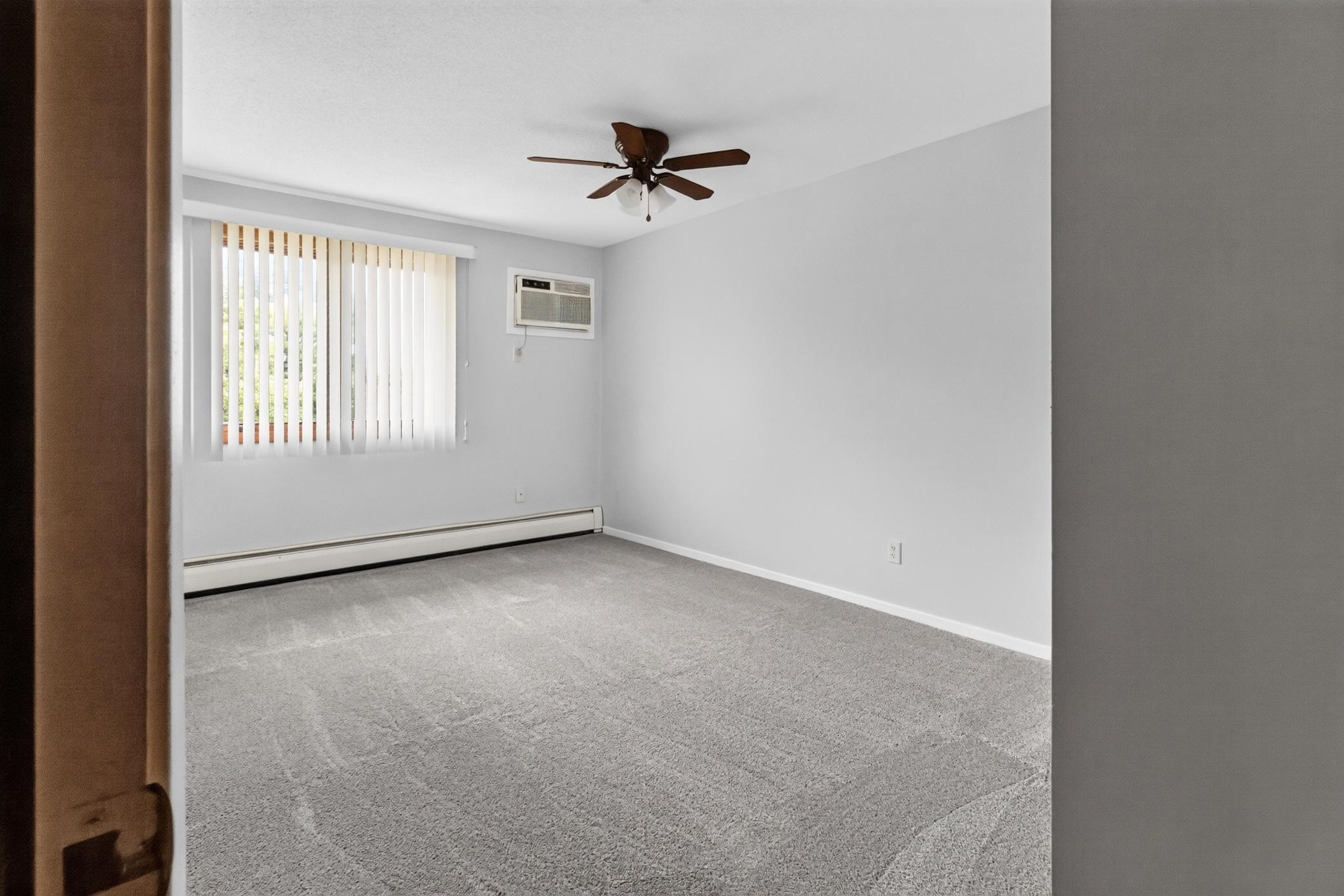 Generously sized bedroom filled with natural light features neutral walls, plush carpeting, a ceiling fan, and a wall A/C unit for localized comfort.