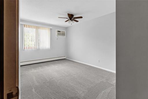 Generously sized bedroom filled with natural light features neutral walls, plush carpeting, a ceiling fan, and a wall A/C unit for localized comfort.