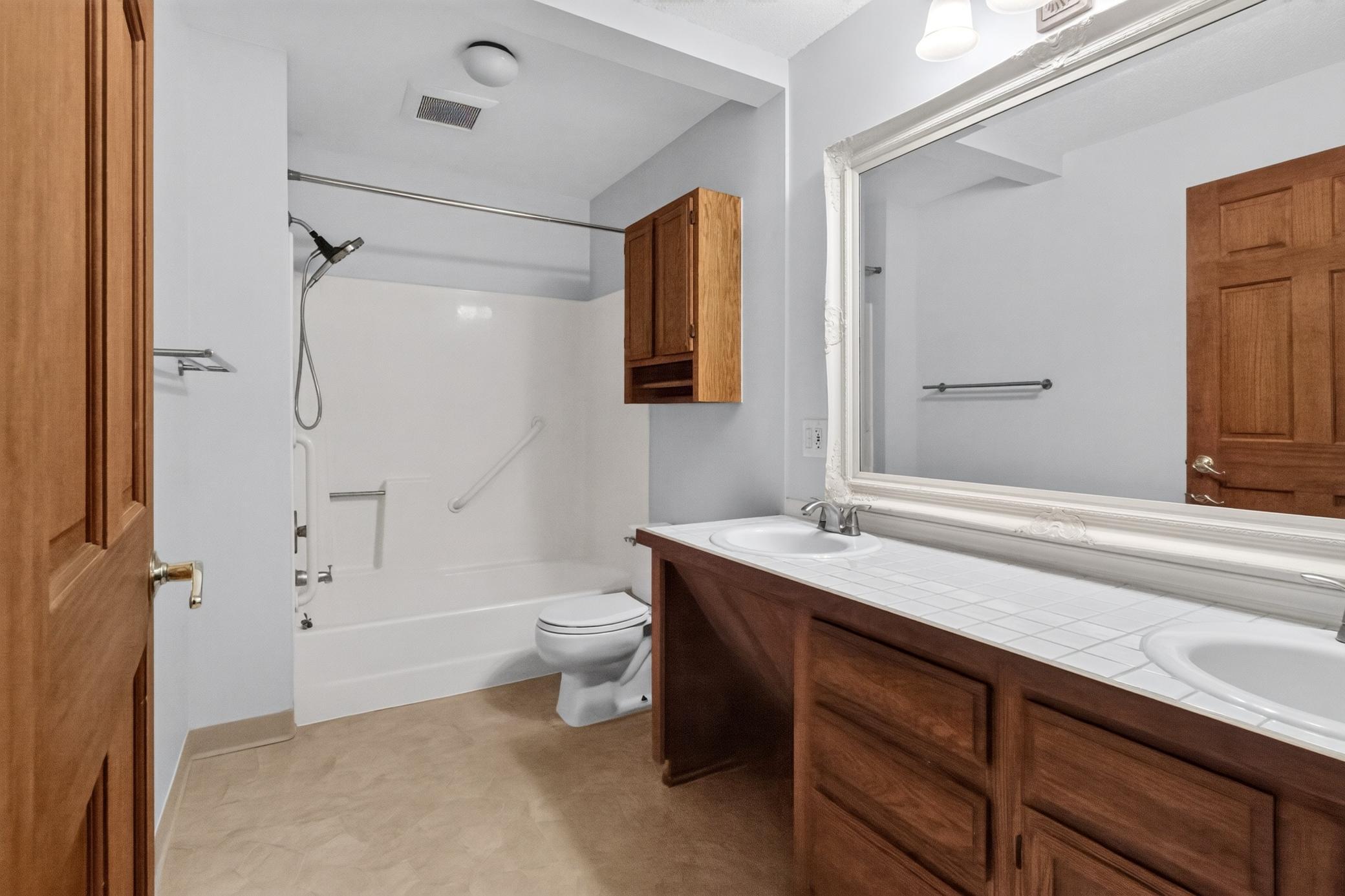 Large full bathroom highlights an expansive double vanity with warm wood tones, a wide framed mirror, and a clean tub/shower combination with integrated safety grab bars.