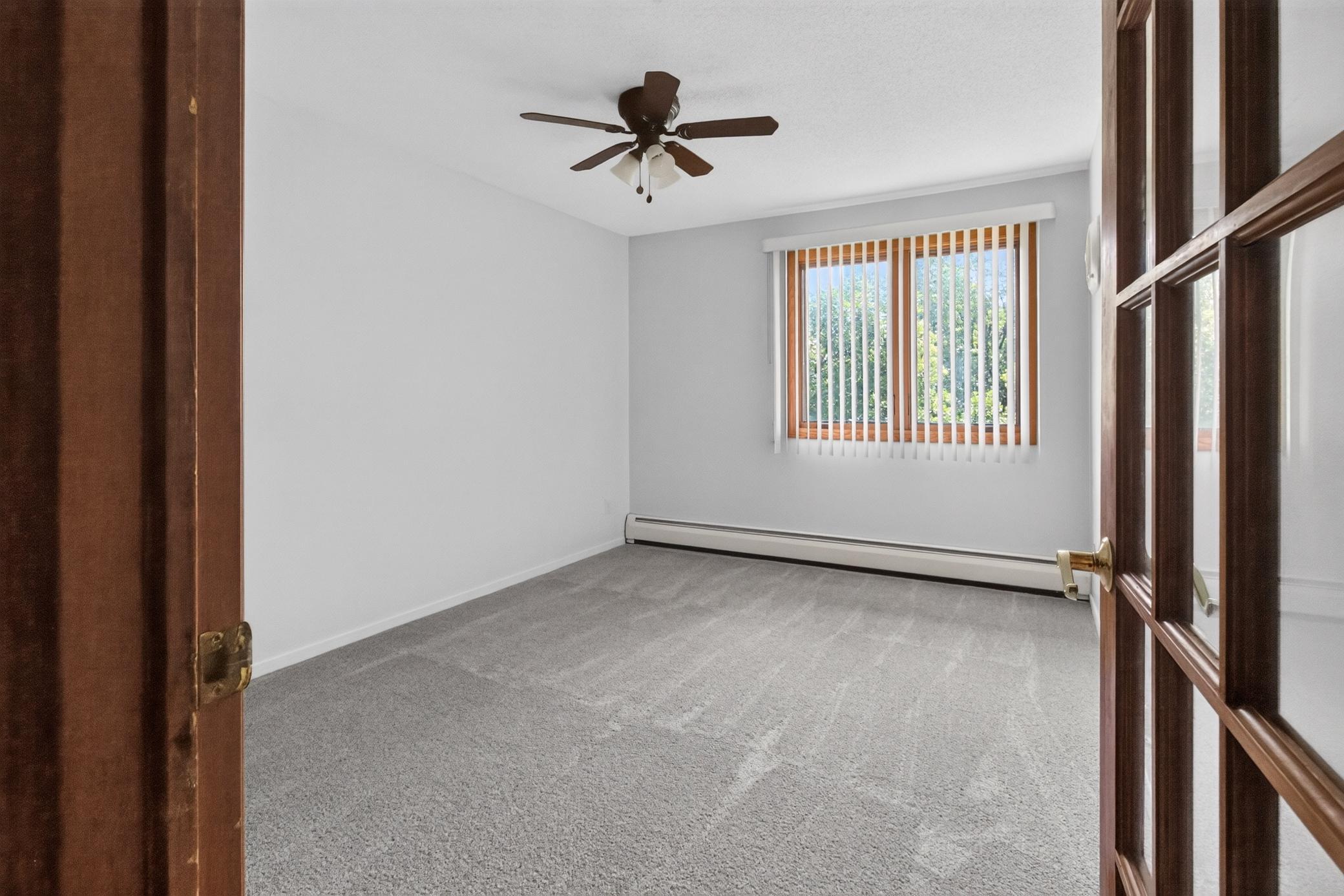 Viewed through elegant glass-paneled French doors, this bright and airy bedroom features soft plush carpeting, neutral walls, a classic ceiling fan, and a large window looking out onto mature greenery.