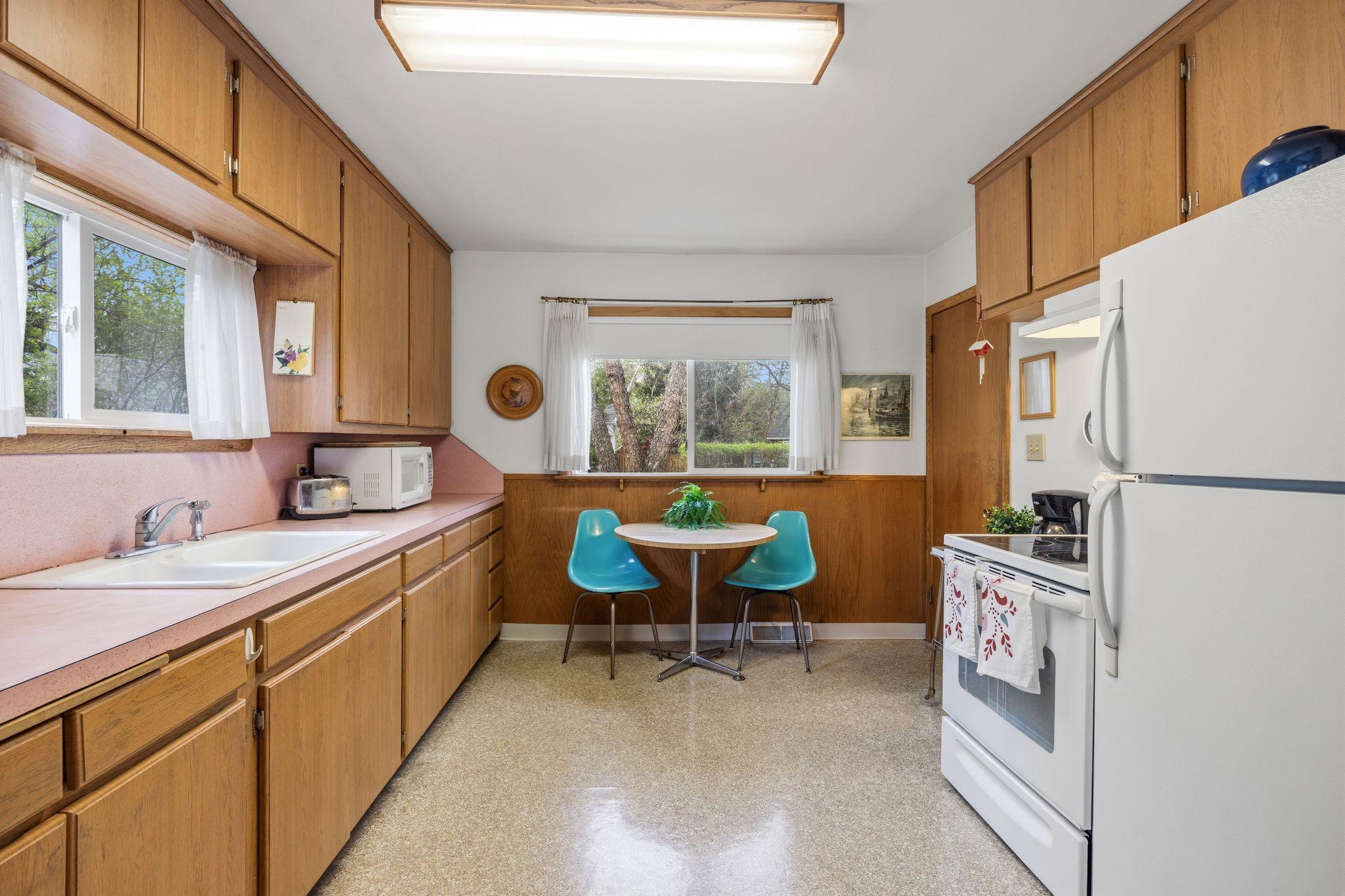 Two large windows flood the kitchen with natural light, highlighting the functional layout and bright atmosphere. The space retains a charming 1950s vibe, anchored by a unique pink countertop.