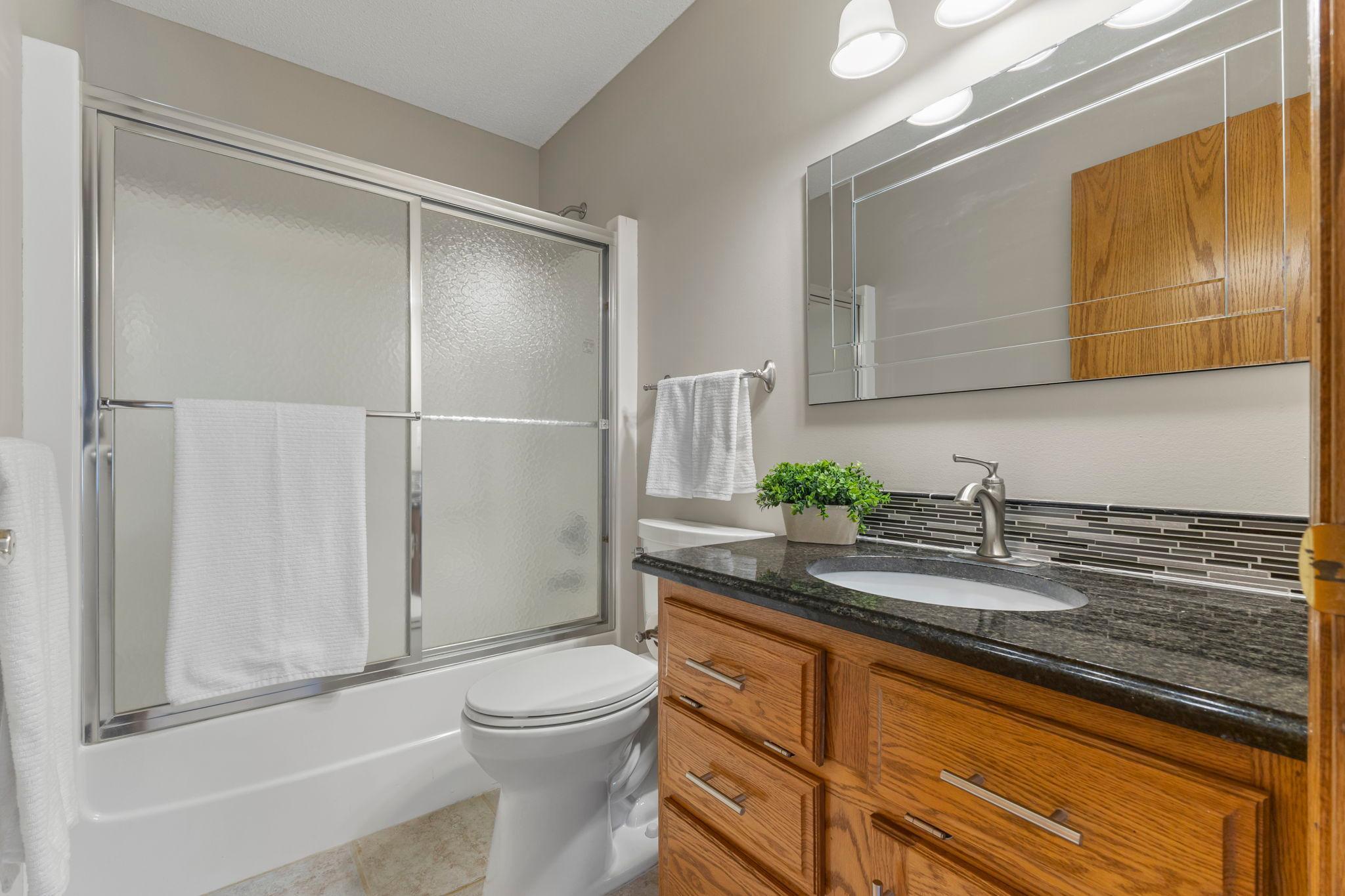 This full bath is perfect for guests and everyday use, featuring a vanity with storage, hard surface countertops, and a tub with glass doors.