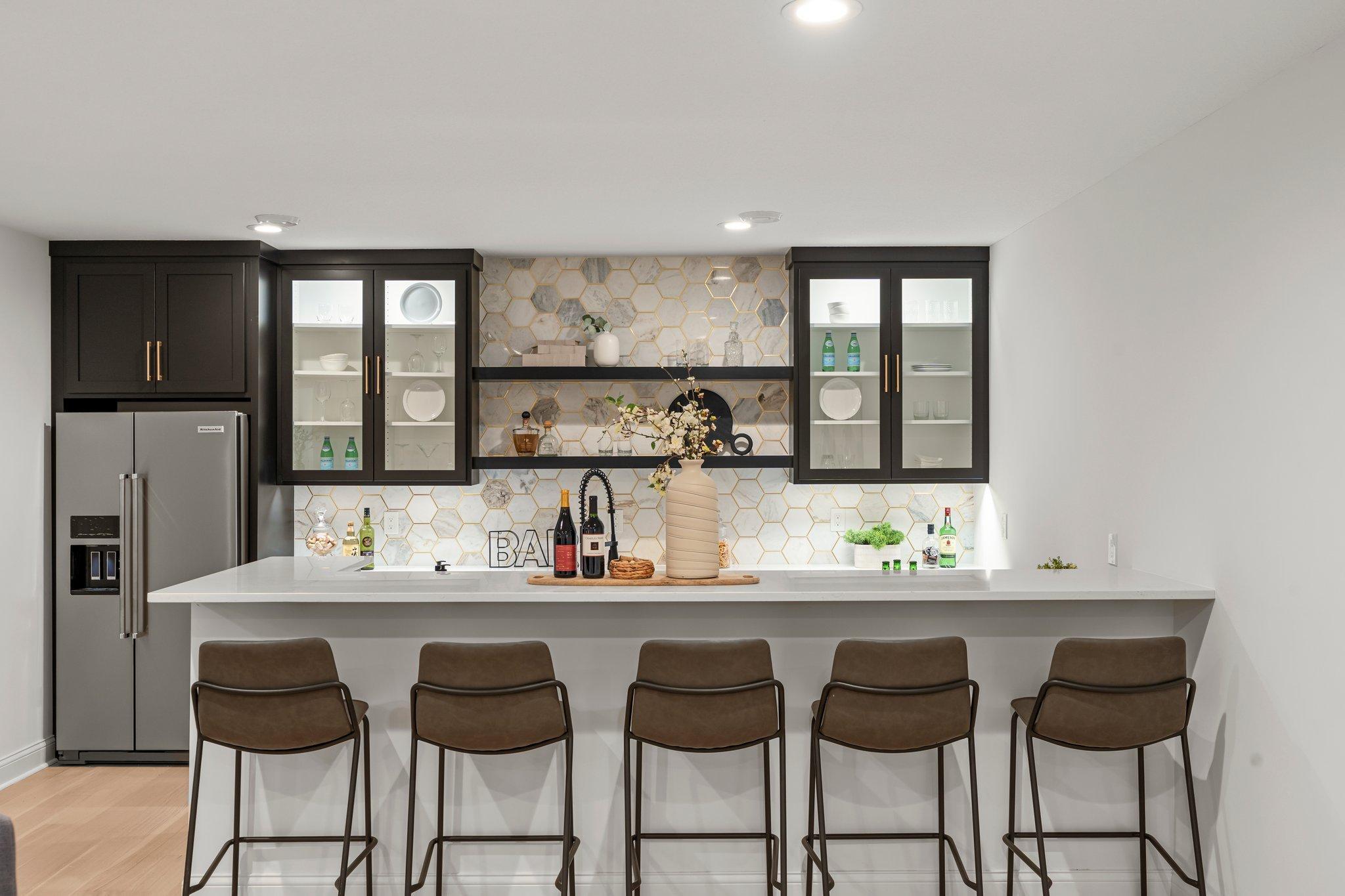 The wet bar area in the lower level features quartz counters, a dishwasher, wine fridge, and a full-size counter-depth refrigerator, all complemented by hexagon tile backsplash that adds a touch of sophistication.