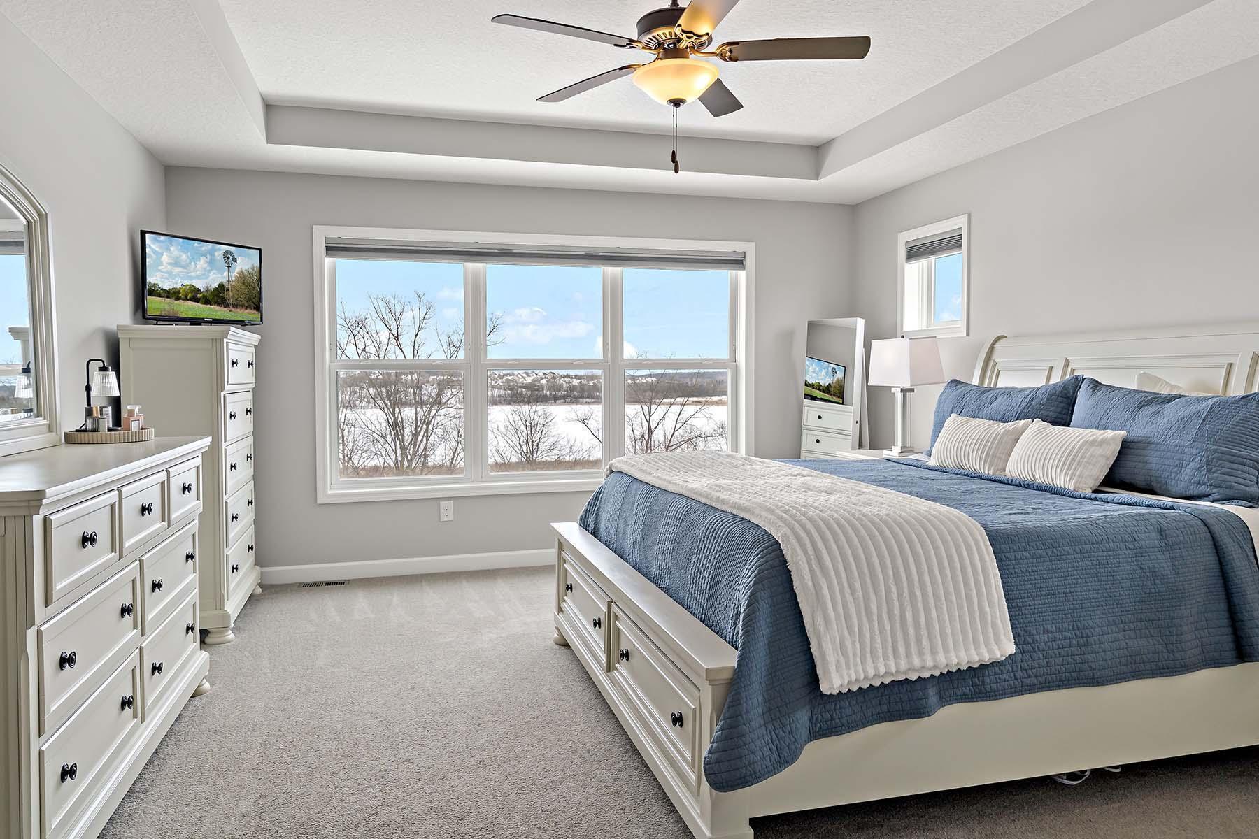 This spacious and serene primary suite features a beautiful tray ceiling and plush carpeting, creating a warm and inviting retreat. A wall of windows fills the room with natural light while showcasing peaceful, scenic views.