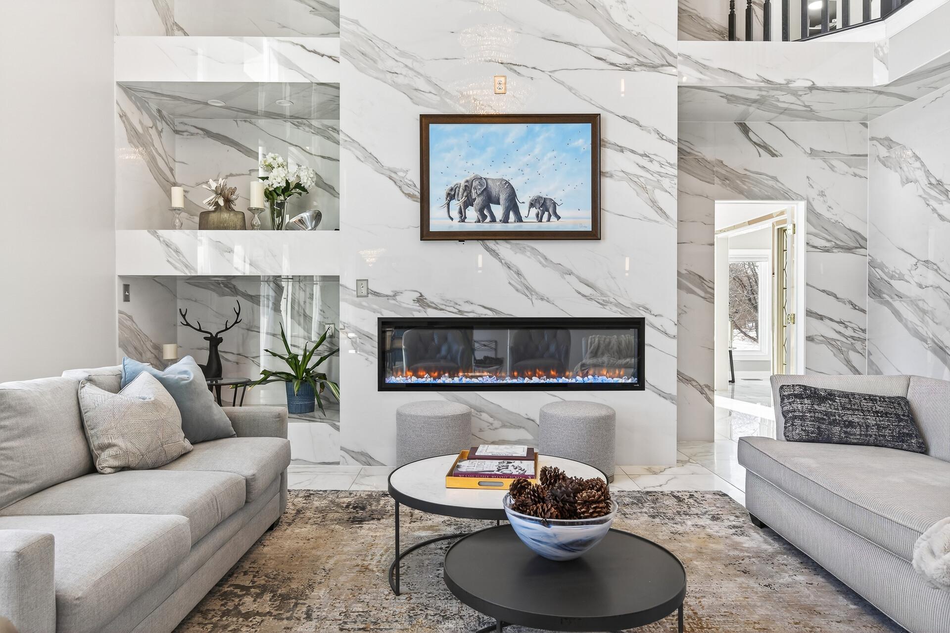 A double-sided electric fireplace, set against a striking marble surround with shelving, anchors the room, while elegant marble flooring and a statement chandelier complete this exceptional space.