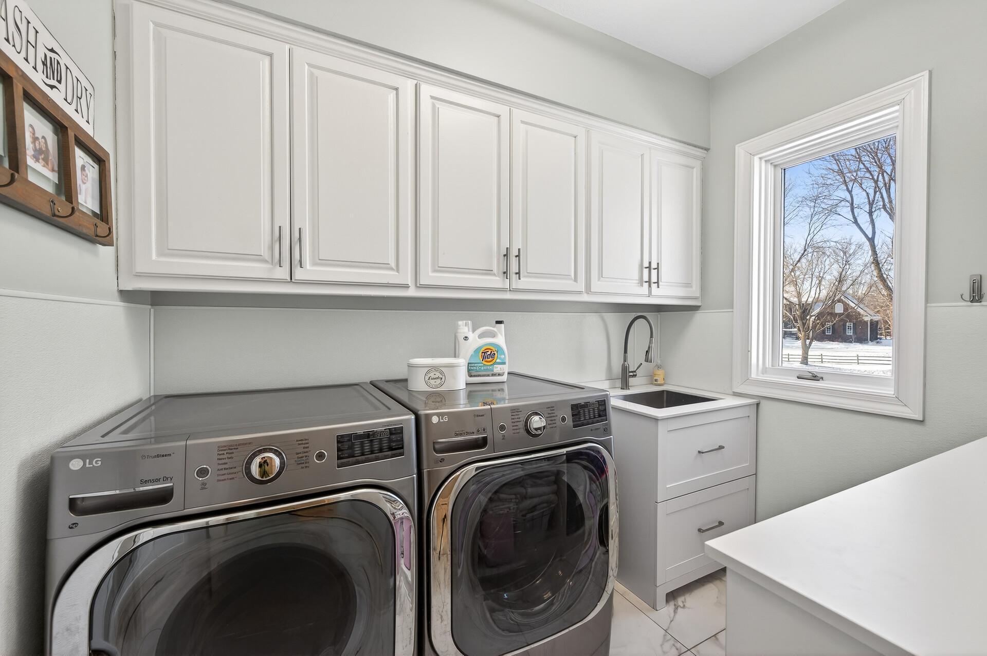 The main-level laundry room is both functional and well-appointed, offering abundant cabinetry and countertop space, a utility sink, front-loading LG washer and dryer, and a built-in fold-down ironing board for added convenience.