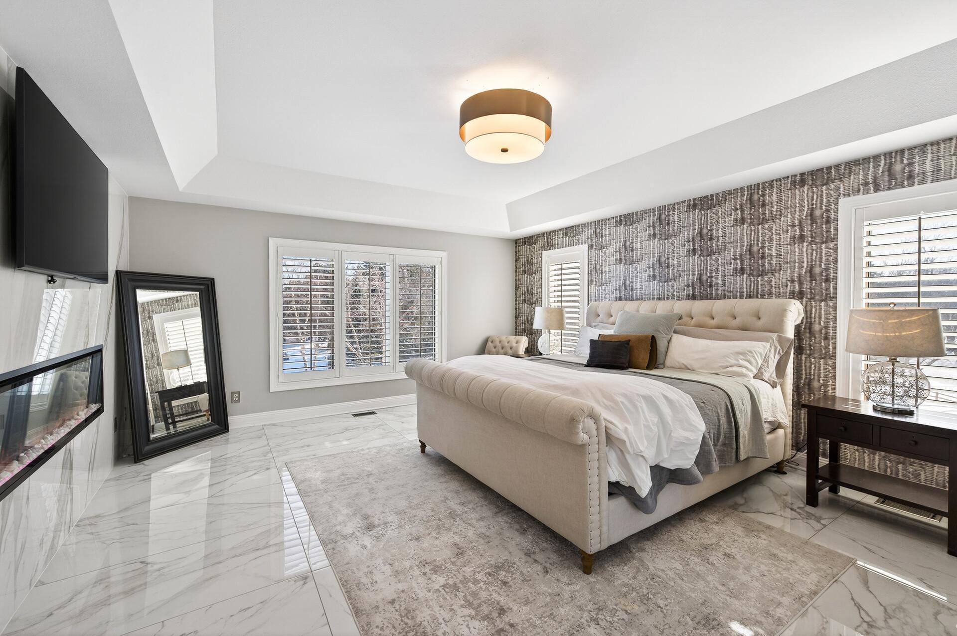 Enter through double doors into a serene retreat featuring tray ceilings, marble flooring, and a cozy electric wall fireplace with a marble surround. Large windows provide abundant natural light, while two generous walk-in closets with built-ins.