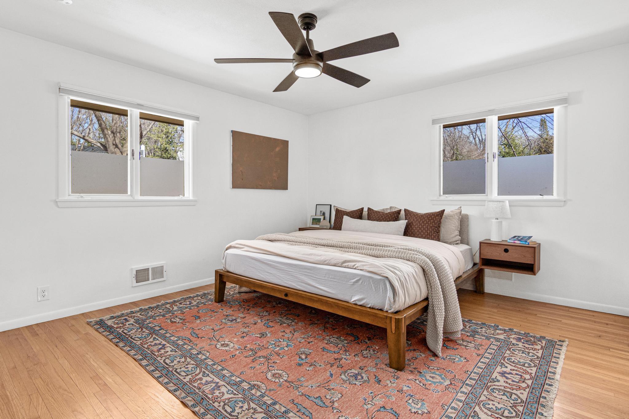 The primary bedroom is a study in calm: white walls, warm hardwood floors, and windows on two sides that fill the room with soft, diffused light filtered through the treetops.