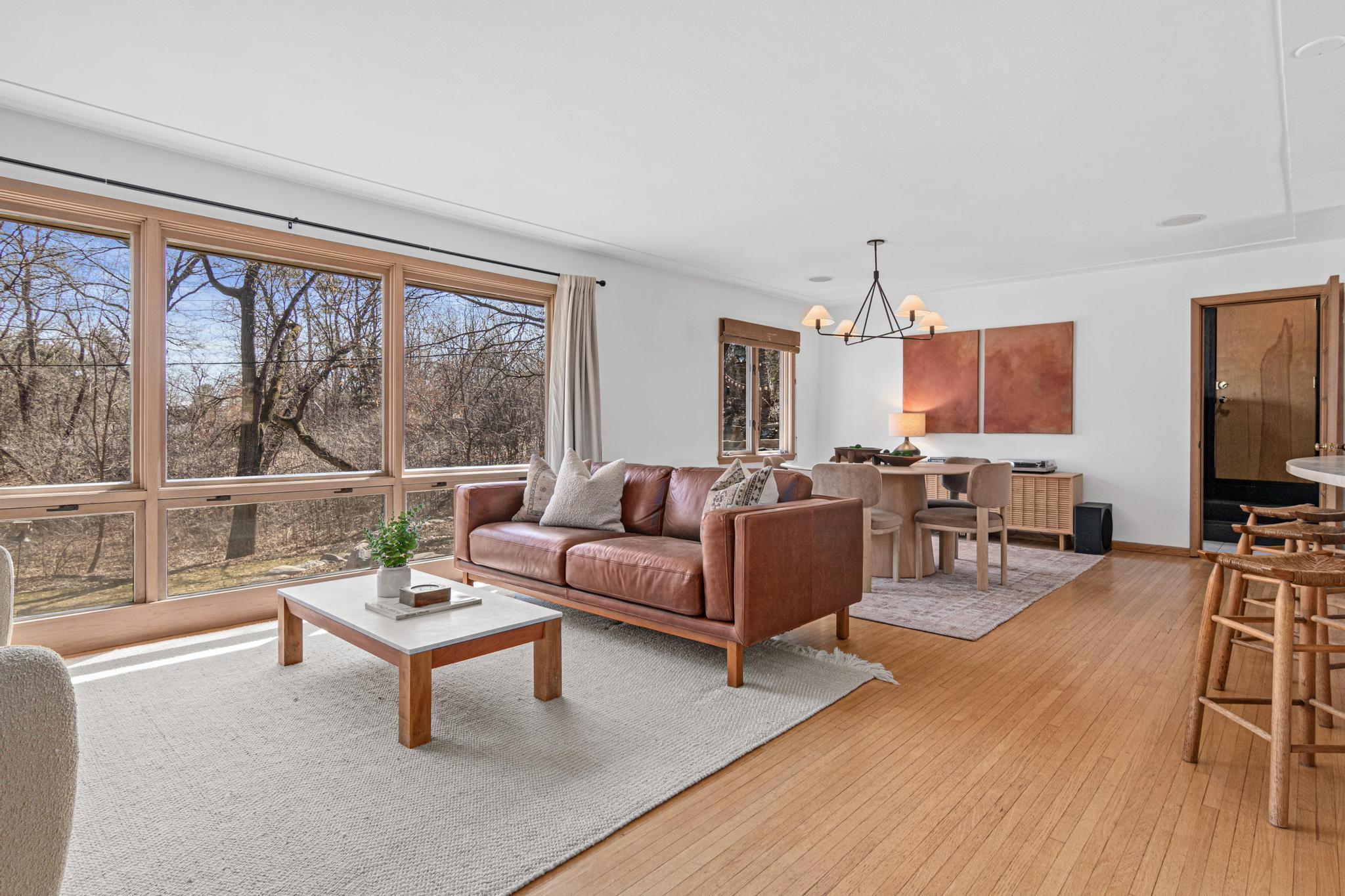 Hardwood floors run continuously through living and dining, a modern chandelier in the dining room, and windows along the entire back wall keep the wooded lot in constant, gorgeous view.
