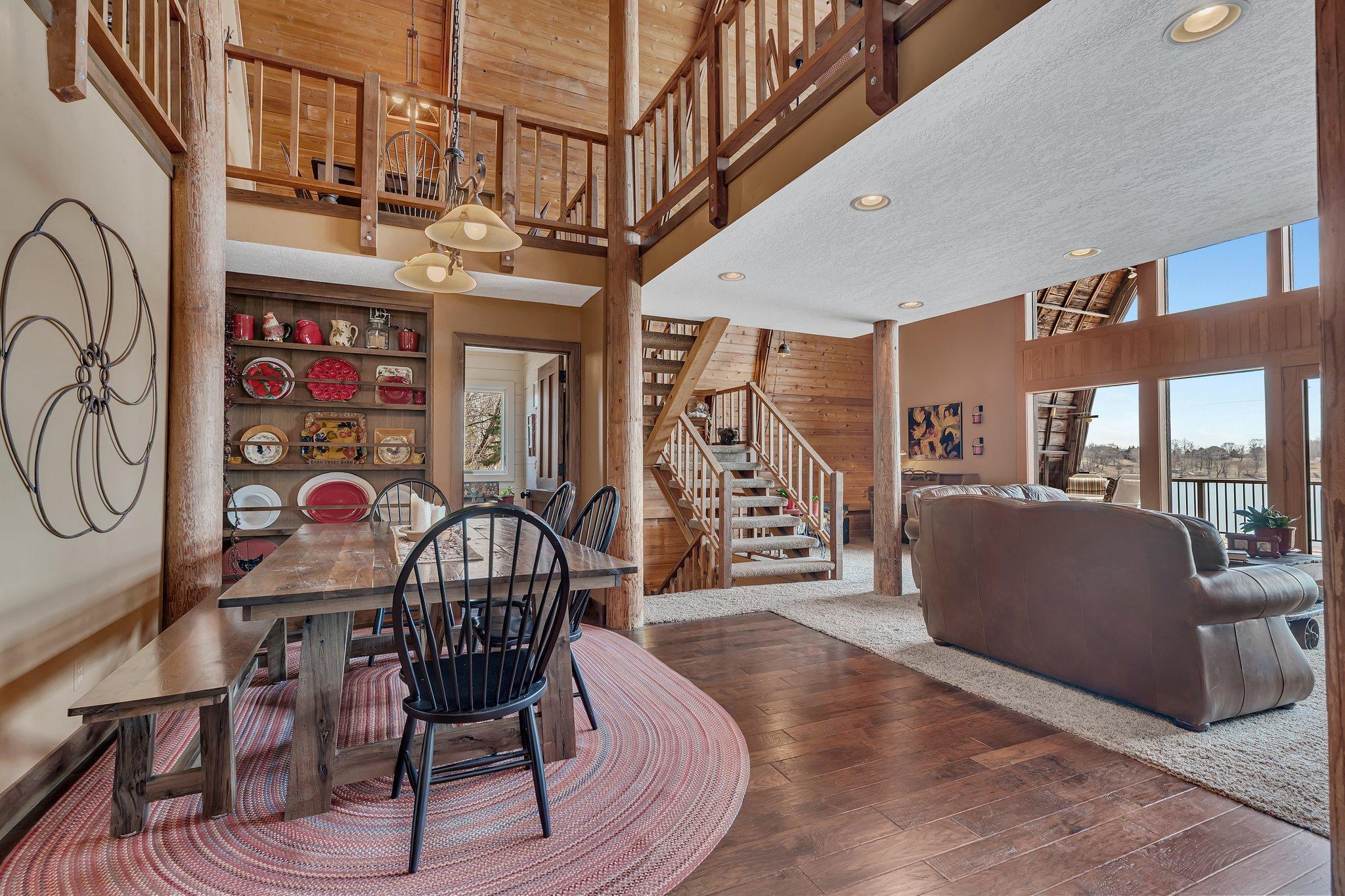 This stunning open-concept interior features a dramatic two-story great room with exposed wooden beams, a loft-style upper level with railing, hardwood floors, and a cozy seating area.