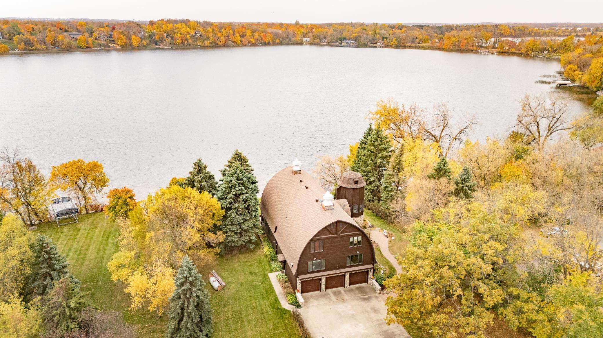 This aerial view showcases a distinctive barrel-vaulted barn home among vibrant fall foliage, and serene Lake Latoka.