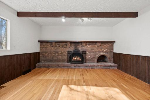 Character-filled main-level family room featuring hardwood floors, rich wood beams, textured walls, a large picture window that fills the space with natural light, and a striking full brick fireplace wall.