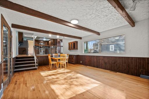 Character-filled main-level family room featuring hardwood floors, rich wood beams, textured walls, a large picture window that fills the space with natural light, and a striking full brick fireplace wall.