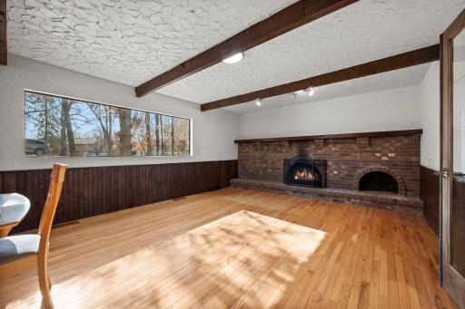 Character-filled main-level family room featuring hardwood floors, rich wood beams, textured walls, a large picture window that fills the space with natural light, and a striking full brick fireplace wall.