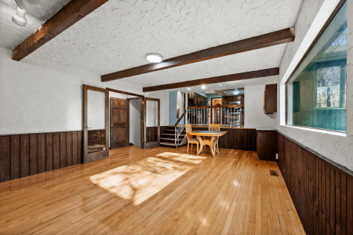 Character-filled main-level family room featuring hardwood floors, rich wood beams, textured walls, a large picture window that fills the space with natural light, and a striking full brick fireplace wall.