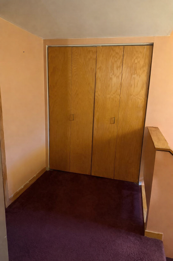 A small hallway landing at the top of the stairs opens to warm peach walls, burgundy carpet, and simple wooden closet doors, creating a quiet, functional pause between levels.