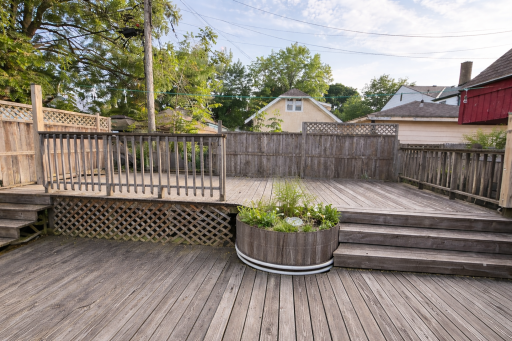 A clean, open multi-level wooden deck with weathered boards and built-in steps is shown in natural daylight, offering a spacious outdoor area framed by privacy fencing and mature trees.