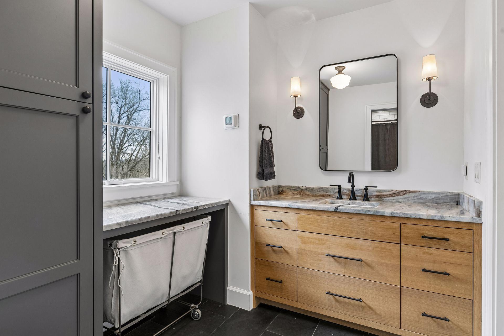 Upper level bath complete with heated slate tile, a modern vanity with mirror, soft wall lights, and natural light from a window. Linen closet and laundry hamper space too!