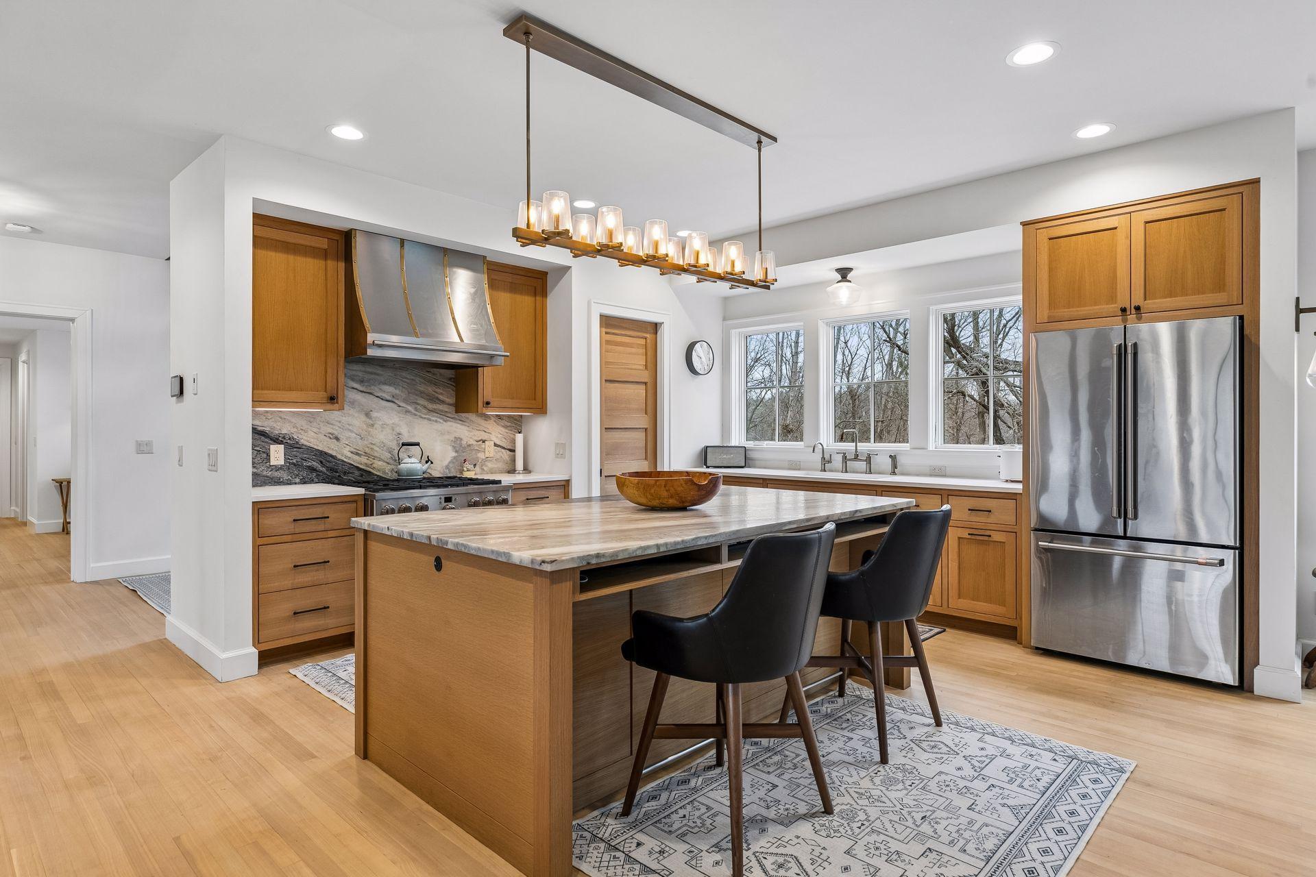 Bright, sophisticated kitchen boasting hardwood floors, a functional island with seating, a large sink framed by three windows, a unique candle-inspired light fixture over the island and recessed lighting for a polished finish.