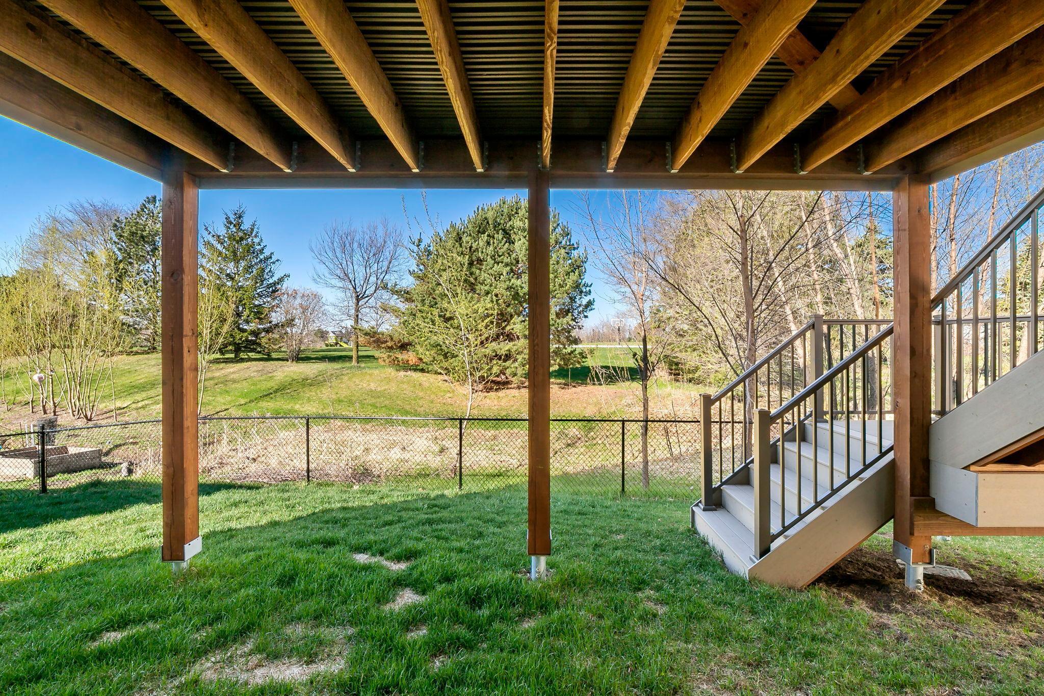 Covered patio area beneath an elevated deck, overlooking a fenced backyard and open green space.
