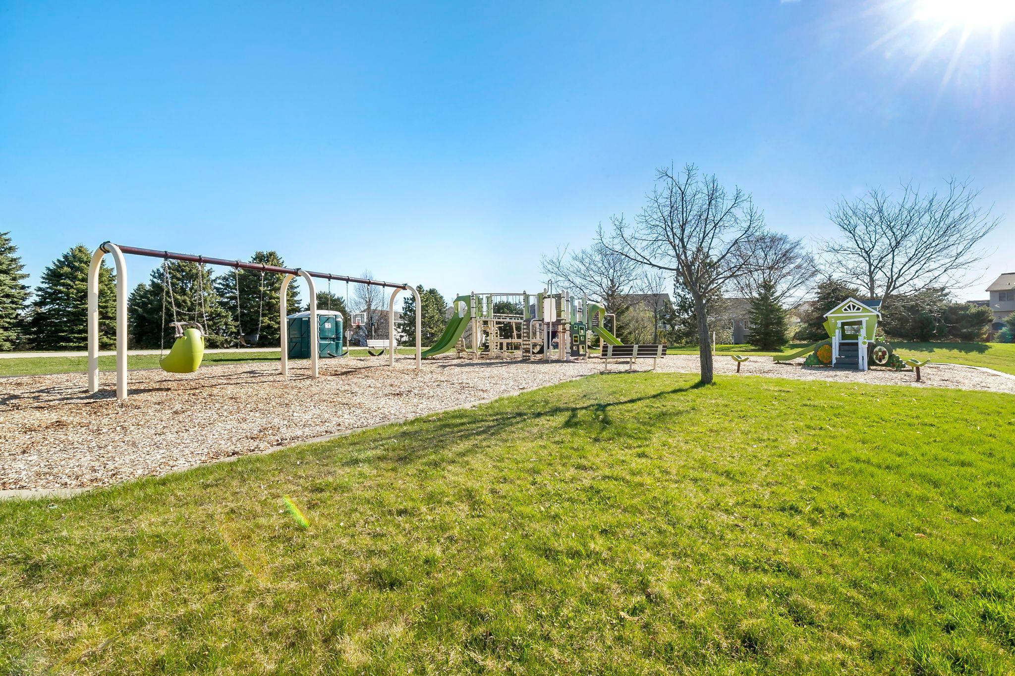 A neighborhood playground with swings, slides, and climbing structures set on a bed of wood chips, surrounded by open green space and nearby homes.