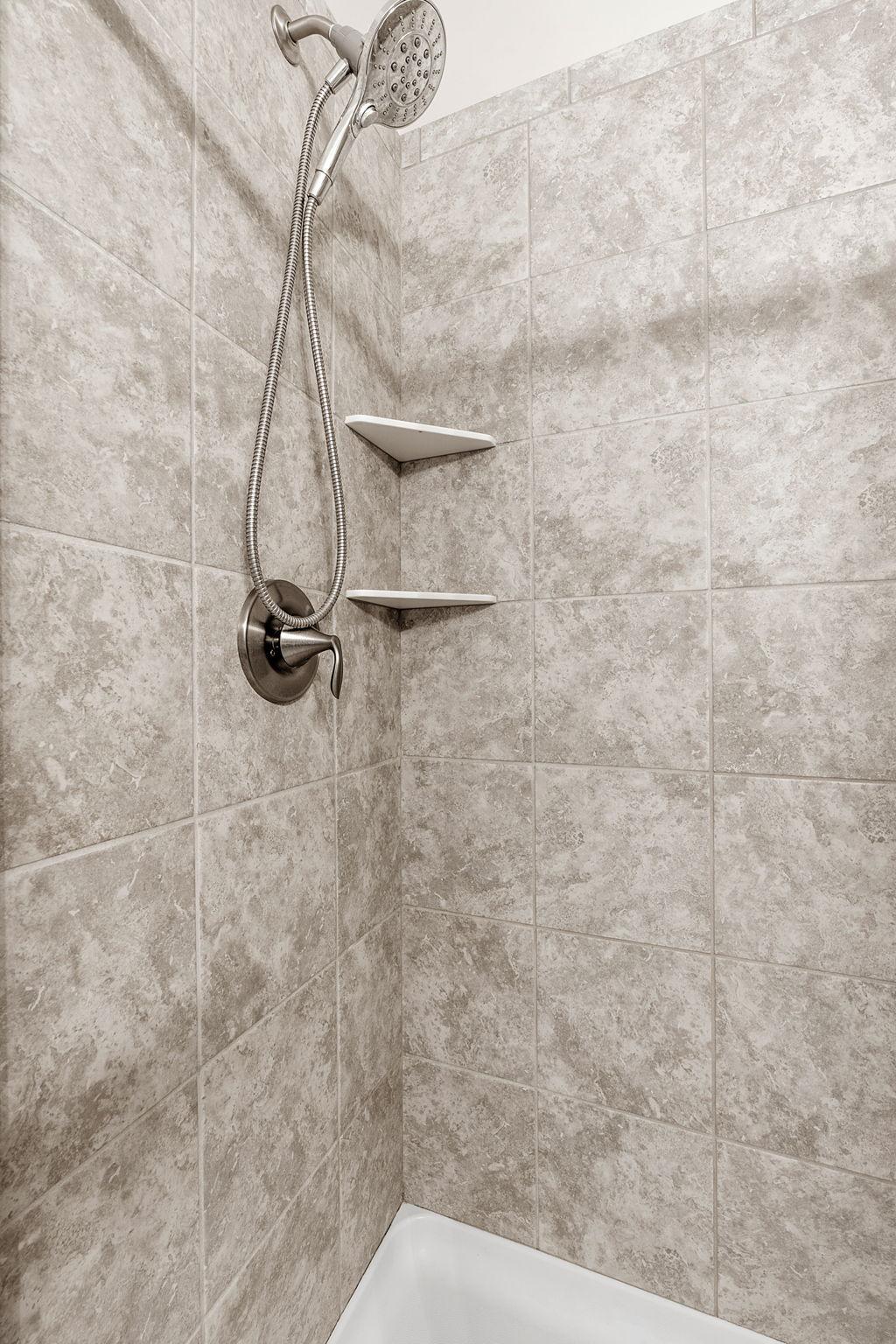 The shower features high-end neutral wall tiling, handy integrated corner shelving, and an adjustable handheld showerhead.