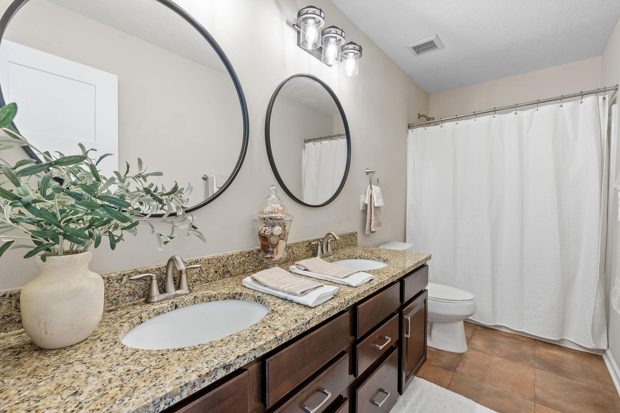 A beautifully appointed guest bathroom boasts a dual-sink granite vanity, rich dark wood cabinetry, modern round mirrors, and a full tub/shower combination.