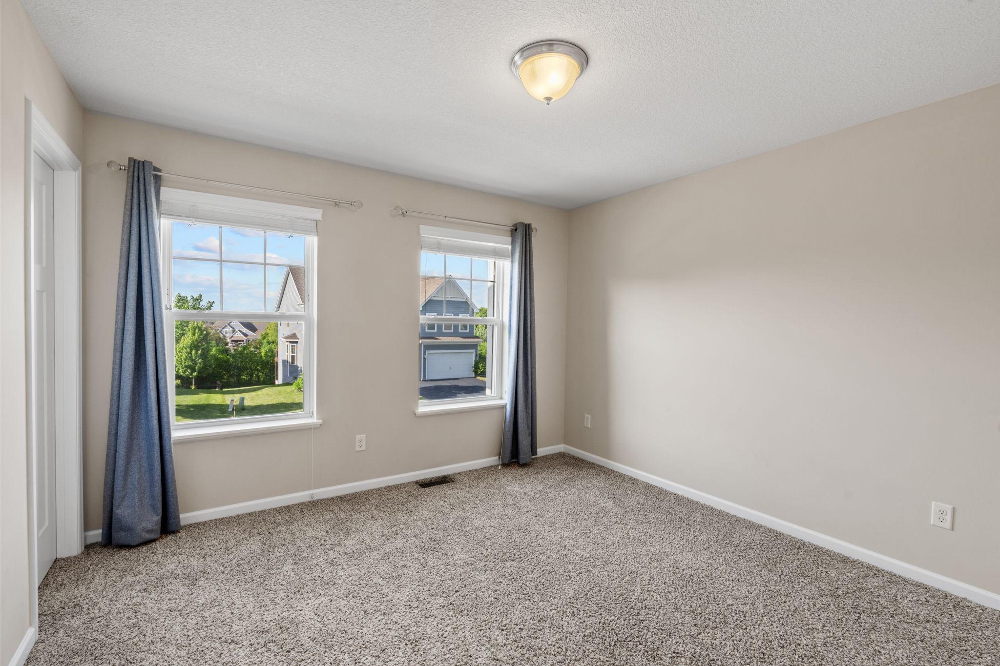This secondary bedroom features soft neutral walls, cozy carpeting, and twin windows that frame lovely neighborhood views.
