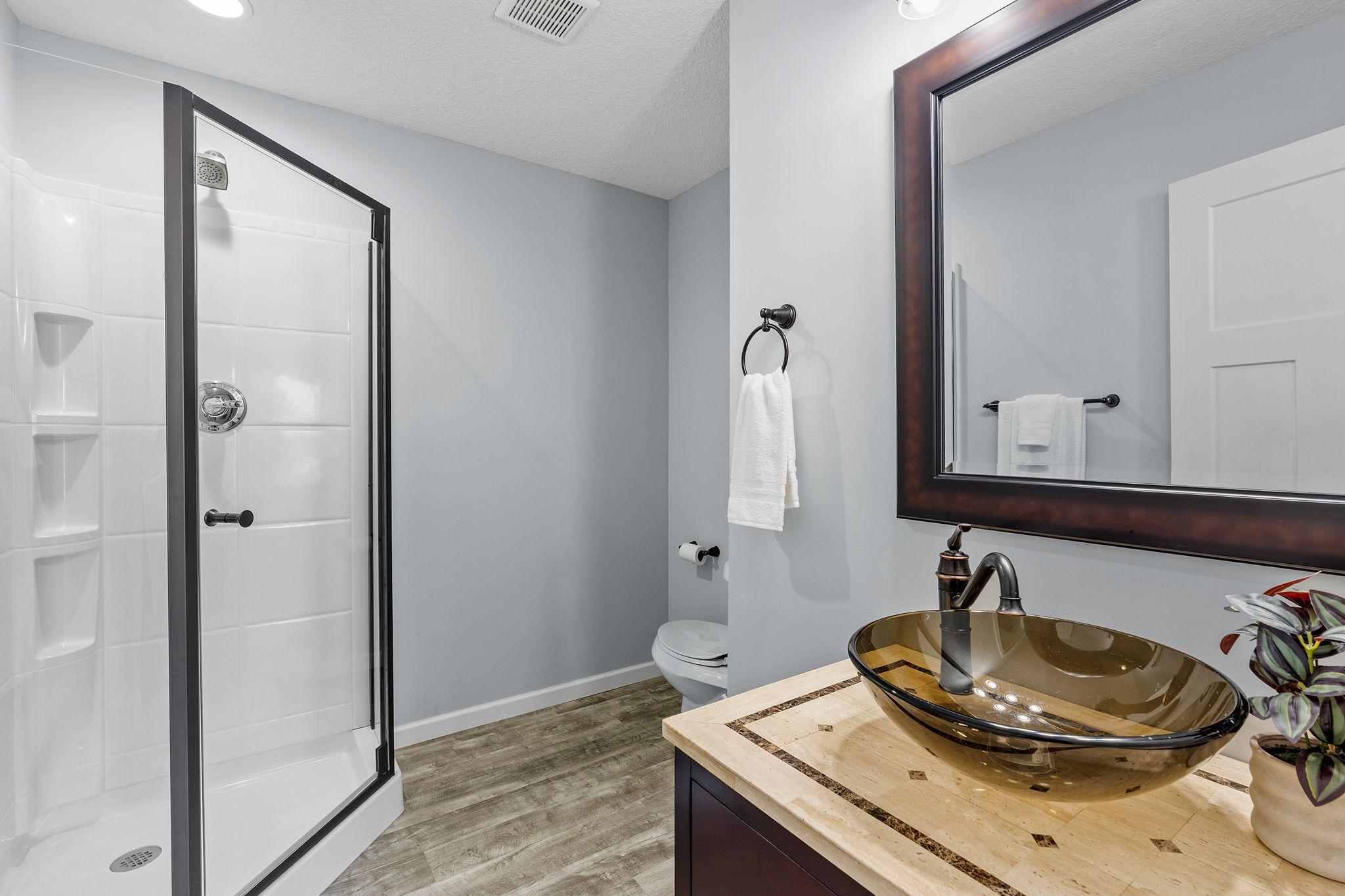 The finished lower level includes a convenient full bath featuring a walk-in shower with integrated shelving, durable wood-look flooring, and a unique glass vessel sink vanity.