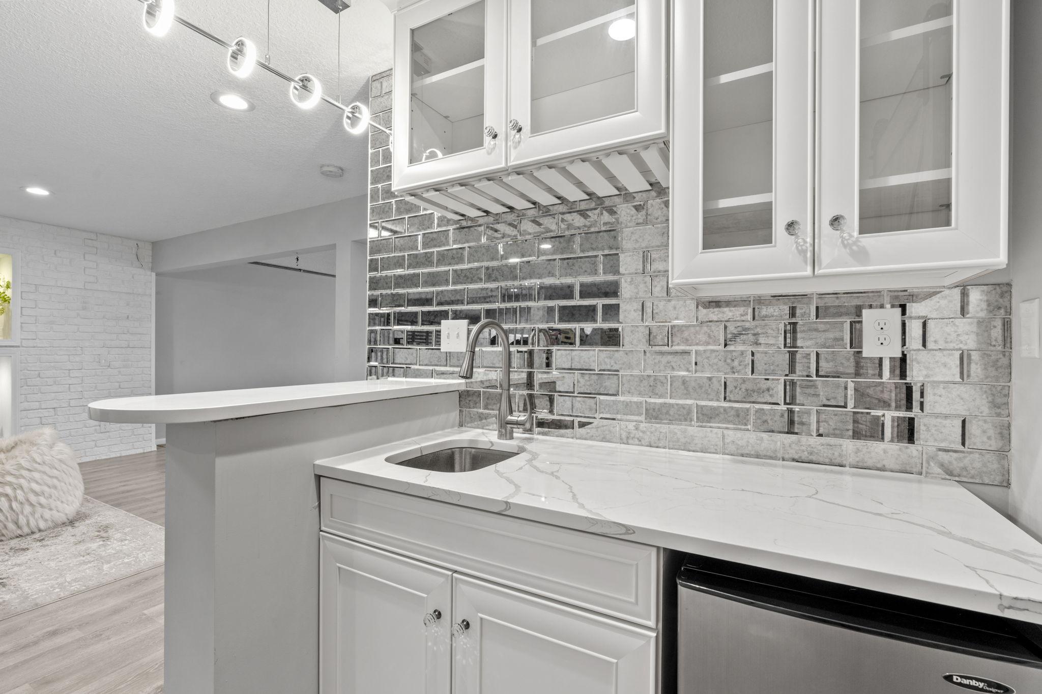 The wet bar boasts a gorgeous veined quartz countertop, an undermount stainless steel sink, and glass-front upper cabinetry complete with integrated wine glass racks.