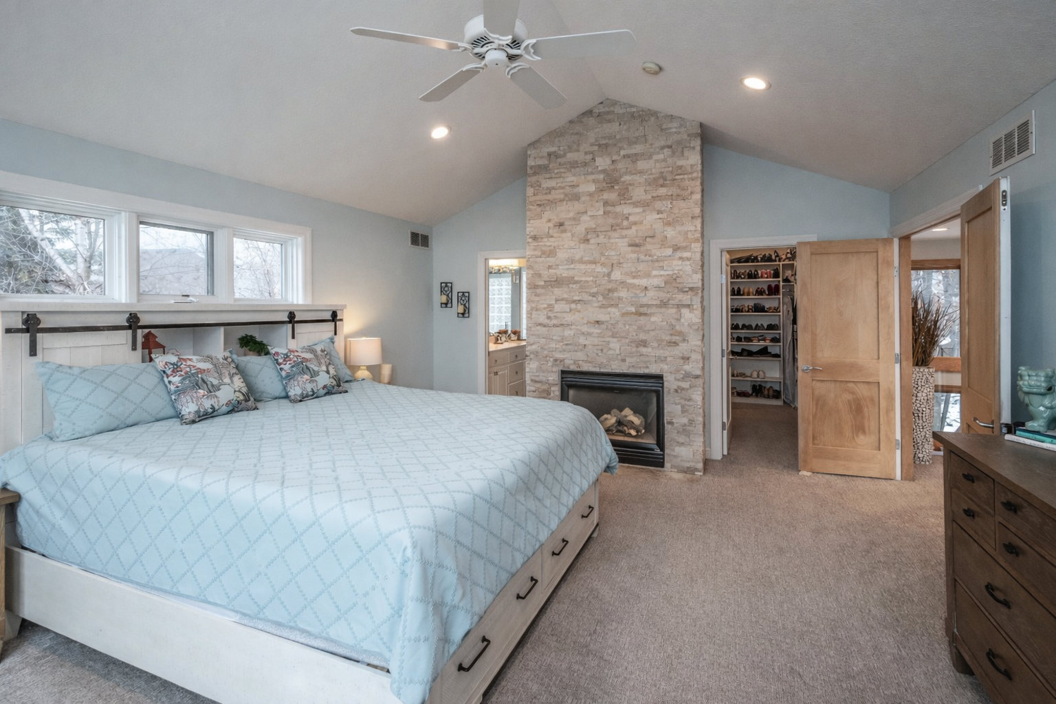 Grand primary bedroom with stacked stone gas fireplace, vaulted ceilings, strategic window placement.