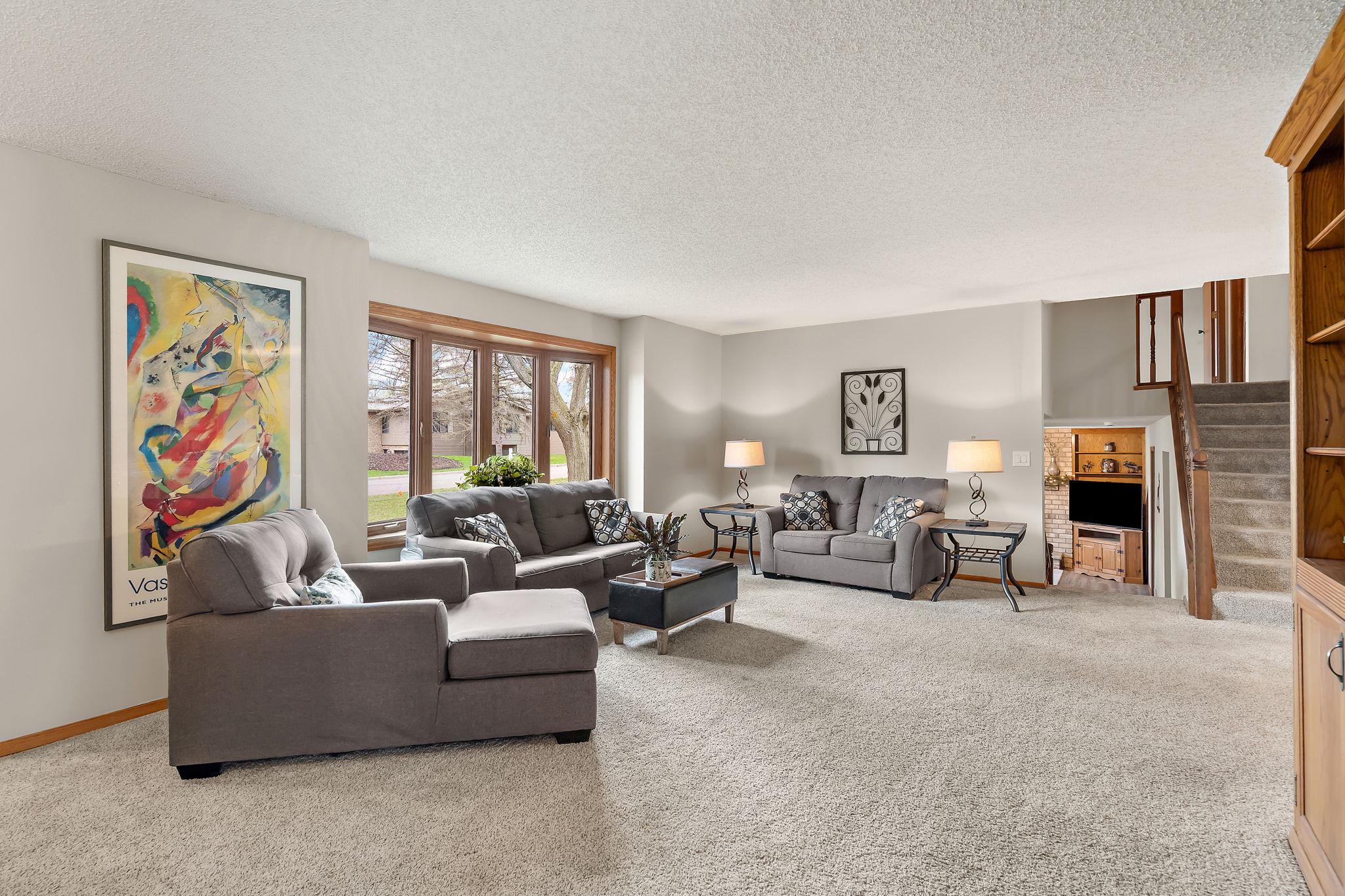 The living room on the main floor features a large bay window facing the street and front yard, allowing natural light to pour into the space. The stairs leading to the upper level and the stairs leading to the lower level are visible beyond.