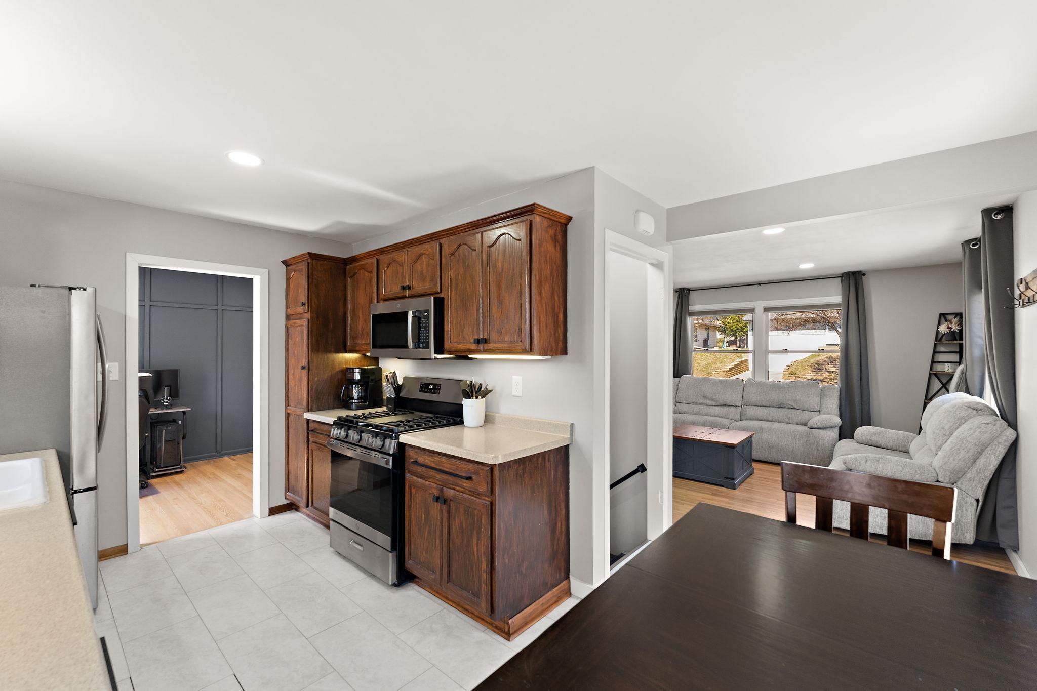 A view from the dining area showing the open kitchen with plenty of cabinet storage and the living room in the room beyond. The hand rail of the stairs leading to the lower level are visible in the open stair way between the kitchen and living room.