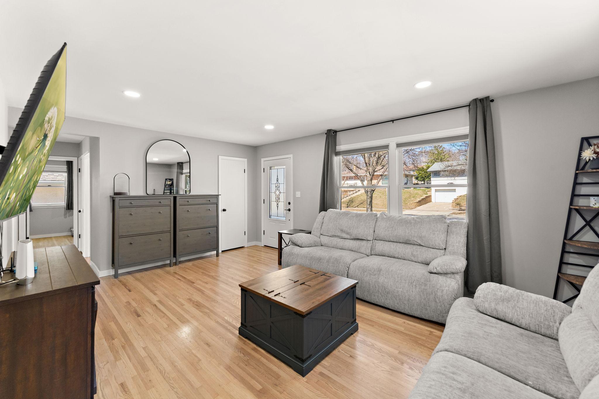 The living room with the front door visible across the room and street view from the large windows. The main floor features newly refinished hardwood floors and fresh interior paint.