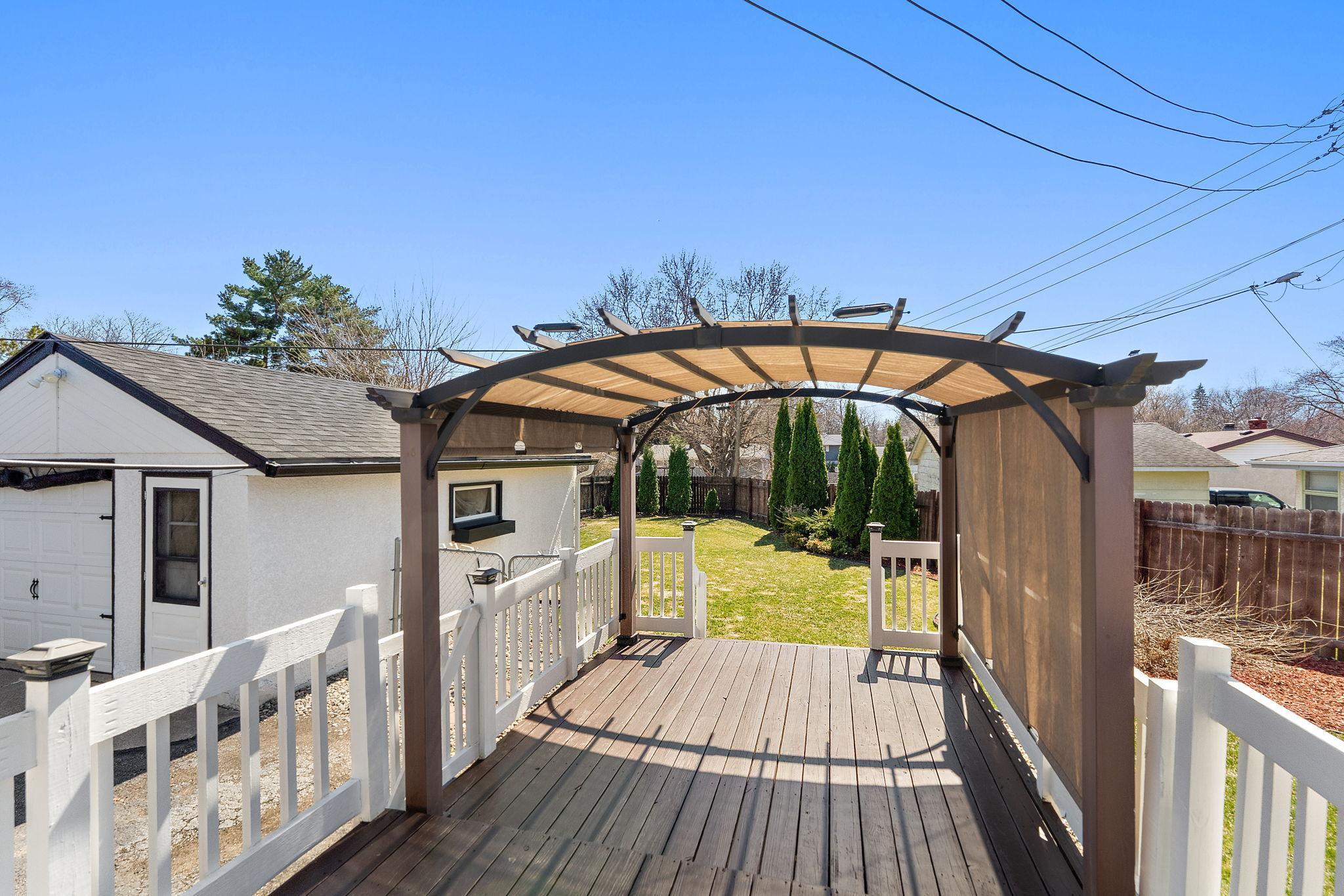 The freshly stained deck has a pergola for shade and a view of the backyard with an access gate to the yard visible on the left in between the garage and deck railing.