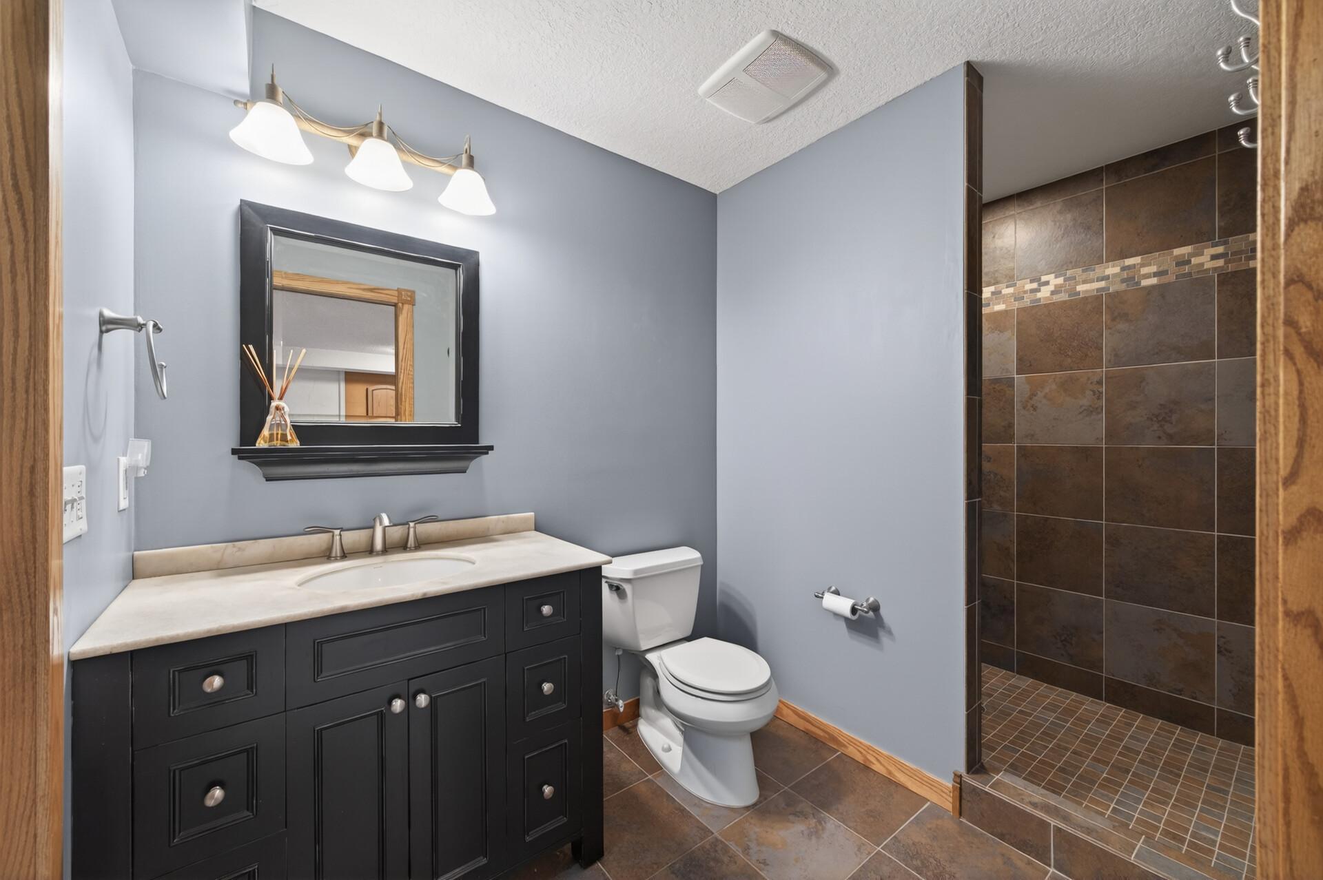The beautifully updated lower level bathroom showcases a spacious walk-in tile shower, offering a spa-like experience with its sleek design and relaxing ambiance. An espresso-colored vanity adds rich contrast, complemented by stylish tile flooring.